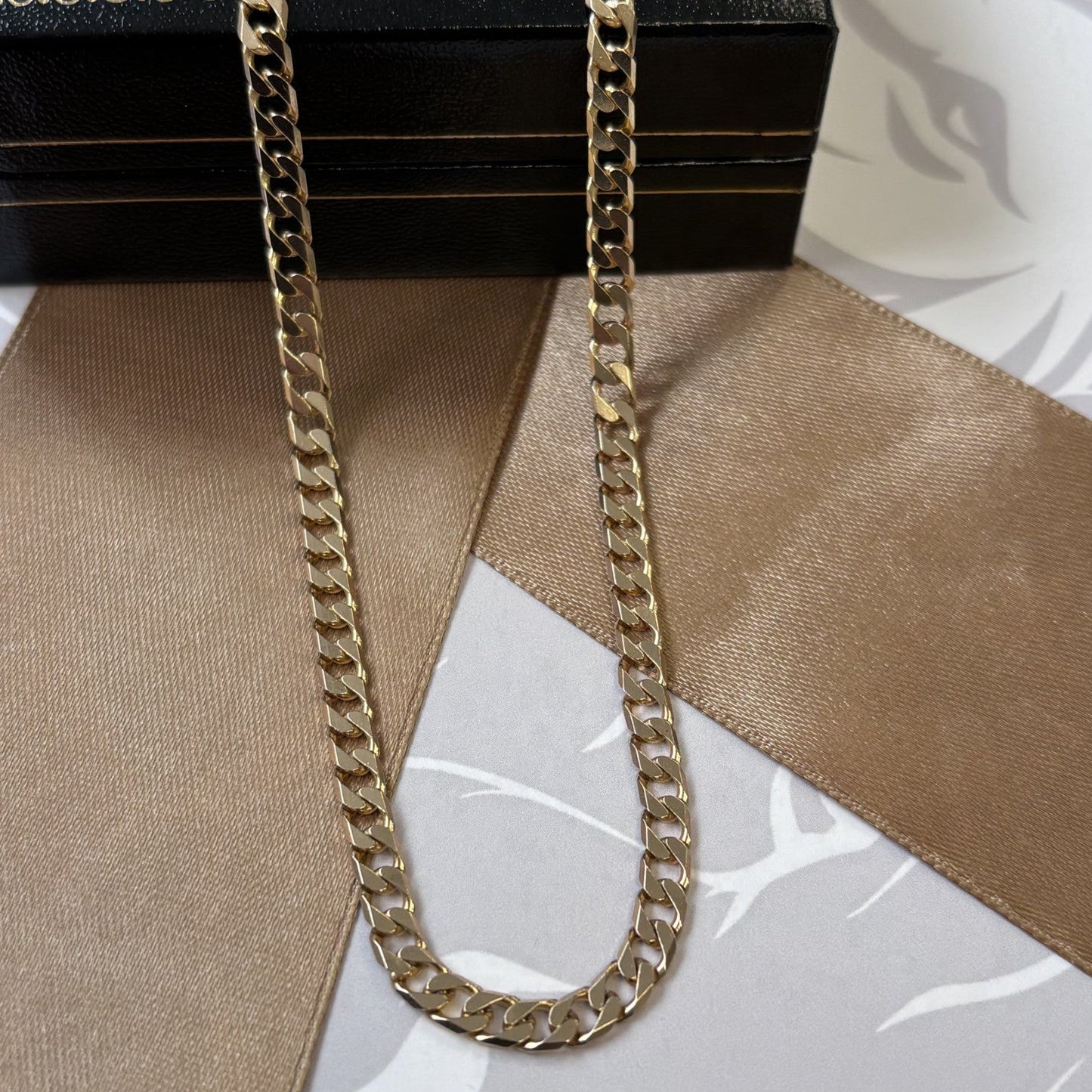 A gold-toned curb chain necklace draped over a black presentation box, with a beige satin ribbon laid across a light gray patterned surface.